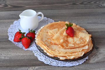 A plate of pancakes, a white jug, and strawberries lie on a wooden table on an openwork napkin. Image with selective focus.