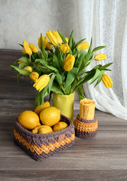 A knitted basket of lemons, a knitted napkin stand, and a vase of yellow tulips sit on a wooden table by the window. Image with selective focus.