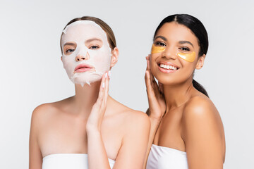 young woman with moisturizing sheet mask near smiling african american friend in eye patches isolated on white
