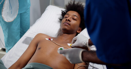 Doctors placing ecg electrodes on young unconscious afro-american patient - obrazy, fototapety, plakaty