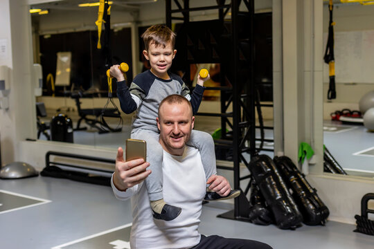 Young father and his adorable son taking selfie in modern fitness center. Young adult father laughs and takes a selfie while his son holds a light yellow weights in his hand. Healthy lifestyle concept