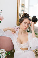 Portrait of a beautiful young girl with long hair in transparent peignoir posing among flowers in a stylish interior. Fresh flowers - roses, tulips. Morning of bride