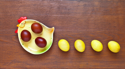 Funny Easter composition with a chicken-shaped plate and painted yellow eggs in a row following a chicken on a wooden background. Blank space for text on top, mock up, copy space, flat lay, close-up