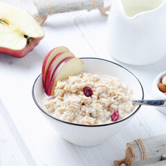Oatmeal porrige with red apple in ceramic bowl over rustic white wooden tray.