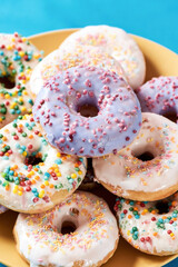 Freshly baked. Close up shot of different colourful round glazed donuts with sprinkles on yellow plate over blue background