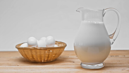 Glass jar with fresh milk near wicker basket with white eggs on wooden table isolated on grey