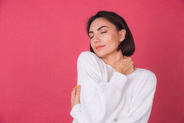 Beautiful woman in casual white sweater on pink red background love concept, holding hands crossed hugging herself isolated copy space closed eyes