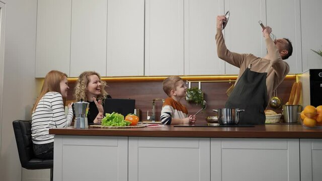 Playful man in apron imagining singing using kitchen devices while his wife and children laughing enjoying amusing performance. Happy deaf parents with kids spending free time in domestic kitchen