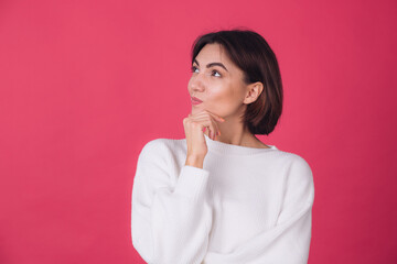 Fototapeta premium Beautiful woman in casual white sweater on pink red background looks to the side thoughtfully with a sweet smile isolated copy space