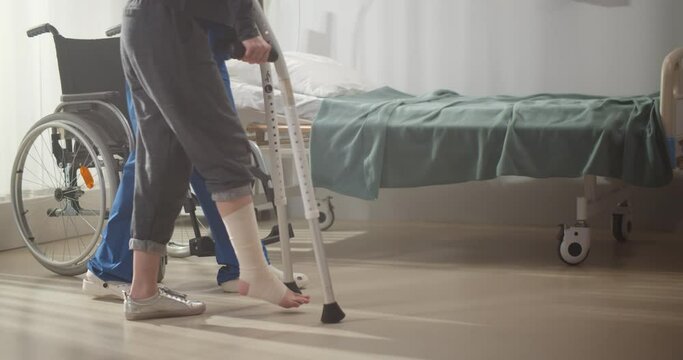 Cropped shot of doctor helping patient with bandaged leg walking on crutches in hospital ward