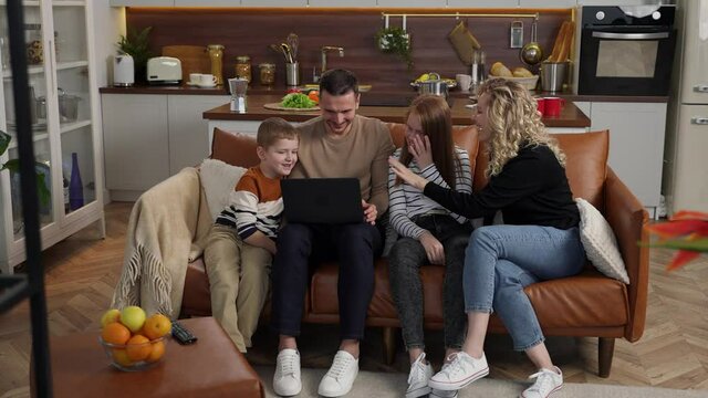 Cheerful hearing impaired couple and children talking with gestures while watching funny video content on laptop. Smiling deaf family enjoying conversation in sign language sitting on sofa at home