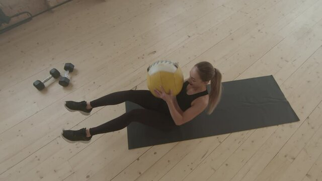 Top-view slowmo of fit woman in tight black sportswear doing side crunches on mat holding heavy weight ball training alone in loft style gym studio
