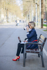 Young woman in a business suit and red shoes drinking takeaway coffee cup and working using laptop on bench in the park, side view. Lonely adult. Female Business Style. Working Freelance