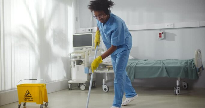 Afro-american male janitor in headphones washing floor in hospital ward