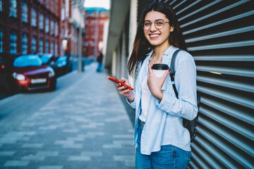 Half length portrait of cheerful woman with coffee to go holding mobile phone smiling at camera at urban settings, funny Caucasian female posing at city street using cellphone technology during travel