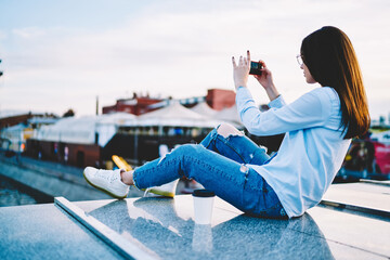 Back view of young woman dressed in casual wear making photo via modern smartphone, millennial...