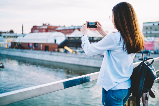 Back view of millennial female tourist focusing mobile camera on river making pictures on smartphone device, casual dressed hipster girl with backpack clicking photos for sharing to travel blog