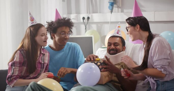 Multiethnic young people congratulating on birthday sick man with neck brace lying in hospital bed