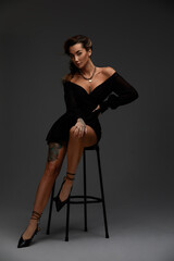 Fototapeta na wymiar Young attractive brunette sexy woman sits on chair in erotic black cocktail dress. Gray background
