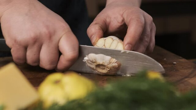 chef in restaurant kitchen cuts garlic with sharp, shiny knife. man prepares food for family and cuts garlic on wooden cutting board.