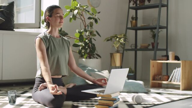 Young woman meditating in lotus pose with hands in mudras on floor in living room while following online yoga class on laptop