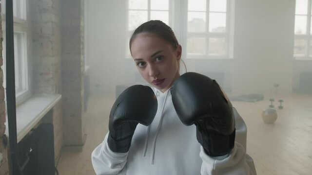 Slow-motion medium portrait of young attractive sportswoman wearing black boxing gloves standing in combat stance looking at camera during indoor boxing training in loft style studio
