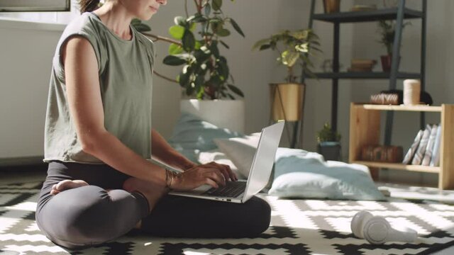 Midsection tracking shot of barefoot woman in sportswear sitting in lotus pose on floor at home and using laptop