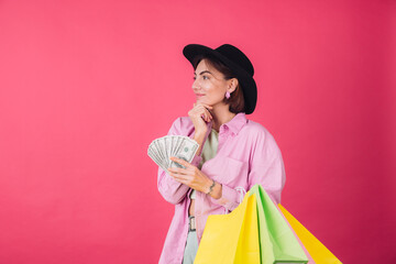 Stylish woman in hat on pink red background isolated space thoughtful hold fan of 100 dollars and shopping bags