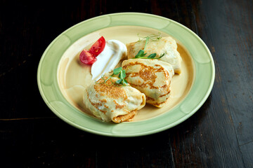 Traditional Ukrainian pancakes with ham and cheese served in a yellow plate with tomatoes and sour cream on a dark background. Ukrainian Blini with filling
