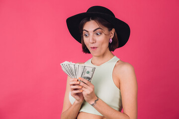 Stylish woman in hat on pink red background isolated space hold fan of 100 dollars money with shocked amazed face mouth open