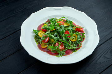 The traditional Italian appetizer salad is vitello tonnato. Thinly sliced beef with rucola, tomatoes and pesto, served in a white plate on a dark background