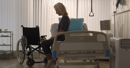 Depressed mature woman patient sitting in hospital bed and looking at wheelchair standing near