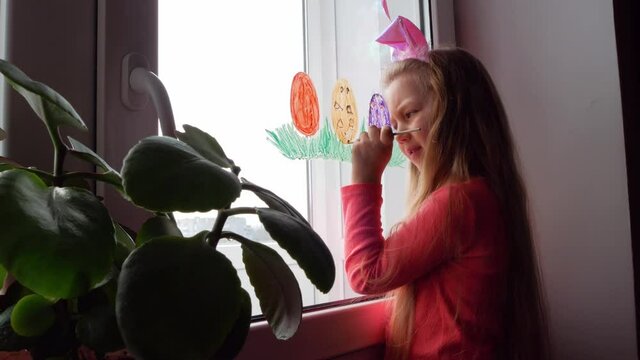 Cute little girl in bunny ears painting Easter eggs with green grass on window at home. Child prepares for spring holiday drawing colorful home decorations.New normal covid-19 lockdown reality concept