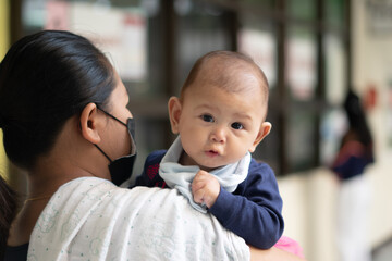 Asian Baby Girl Looking Straight and tired after vaccine shot while being held by mother wearing a mask during pandemic