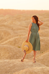 Young beautiful smiling woman with long hair in blue dress with straw hat in hand standing on sand...