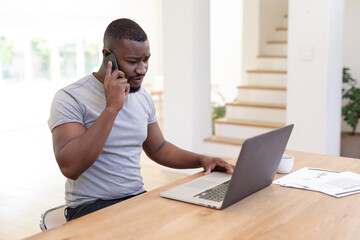 Obraz premium African american man working from home sitting at table using laptop and talking on smartphone,