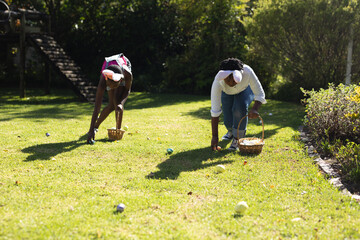 African american brother and sister wearing bunny ears easter egg hunting in garden