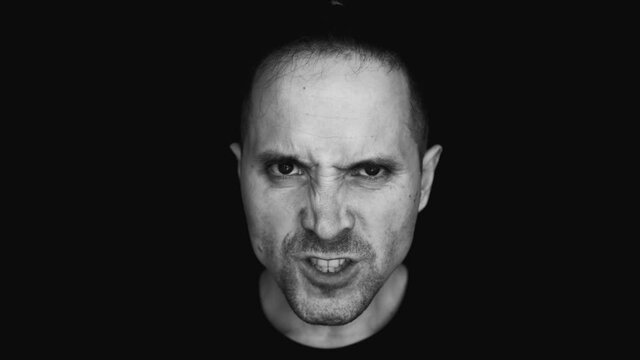 Aggression. A black-and-white photo of an angry man. He grins. The eyebrows are drawn together. Look under your eyebrows. Discontent. Anger. Black background.