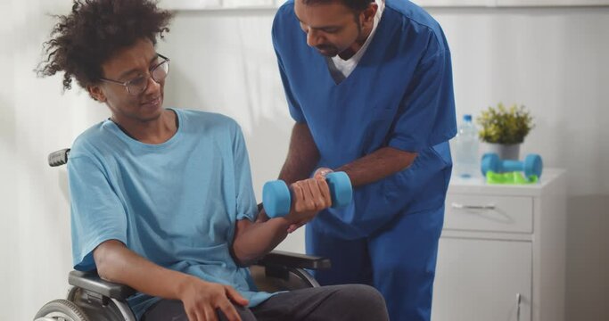 Indian physiotherapist helping disabled young man in wheelchair to work out with dumbbells