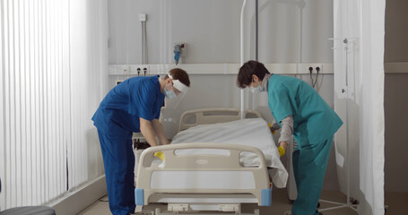 Medical workers in safety mask and gloves changing bed sheets in hospital room