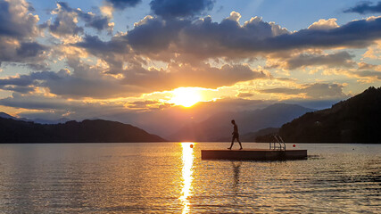 Fototapeta na wymiar A man running along wooden platform drifting on the MIllstaetter lake during the sunset. Jumping into deep water. The sun sets behind high Alps. Calm surface of the lake reflects the orange sky. Fun