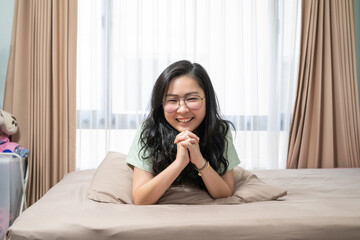 Beautiful Asian woman hold her hands together while lie on her stomach on bed in bedroom with a pastel green - brown color theme.