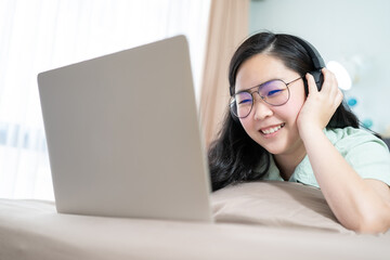 Beautiful Asian woman is using a headset with laptop and lying on the bed in her bedroom with a pastel green - brown color theme.