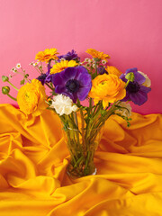 Bouquet of spring flowers: purple anemone flowers and yellow ranunculus in glass vase on yellow draped textile background. Creative, vintage floral concept. Minimalism aesthetic. 