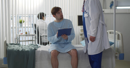 Sad young male patient signing medical form sitting on hospital bed in modern clinic.