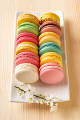Multicolored macaroons and spring cherry blossom flowers on a white rectangular plate against natupal wood table. Sweet breakfast with delicious almond meringue cookies. Colorful macaron cake dessert.