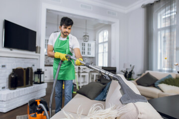 Fototapeta na wymiar young indian man removing dust from sofa with vacuum cleaner