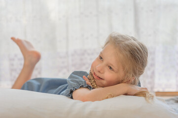 Fototapeta premium a little girl with two pigtails of blond hair lies on her stomach on the floor of the house and shows her tongue