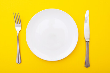 Ceramic white plate with knife and fork on yellow surface, kitchen table, lay out, top view, space for text