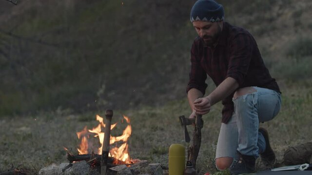 Evening at hiker camp. Traveller throwing stick in fire, chopping wood to start campfire, holding an axe, prepare for the night on hiking adventure in woods, making bonfire